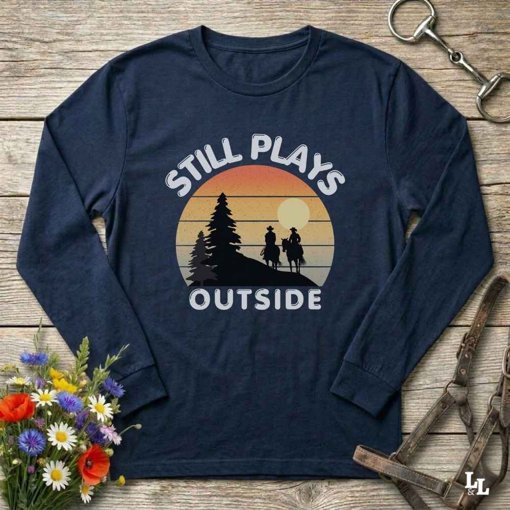A navy blue long-sleeve t-shirt with a retro sunset graphic and the text 'STILL PLAYS OUTSIDE' on the front, styled in a flat-lay on a wooden background with equestrian accessories and flowers.