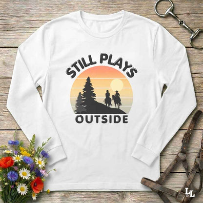 A white long-sleeve t-shirt with a retro-style graphic print of a sunset and horse riders, featuring the text 'STILL PLAYS OUTSIDE'. The shirt is displayed in a flat-lay arrangement on a rustic wooden background, styled with a small bouquet of colorful wildflowers, a metal horse bit, and a brown leather strap.