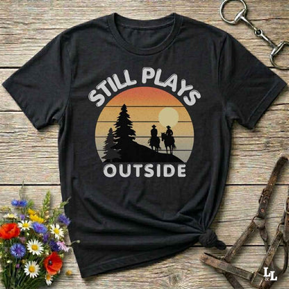 A black short-sleeve graphic T-shirt laid flat on a rustic wooden surface, with a sunset-style circular print showing two horse riders and the words “STILL PLAYS OUTSIDE.” Decorative flowers and equestrian gear appear around the shirt.