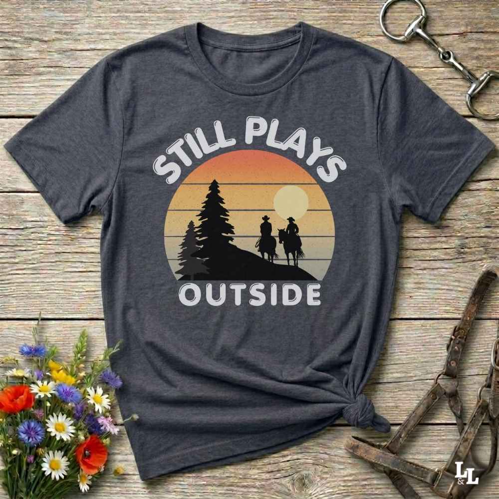 A dark charcoal gray t-shirt with a retro sunset graphic and the text 'STILL PLAYS OUTSIDE' in white. The shirt is styled in a flat-lay on a rustic wooden background with equestrian accessories (bridle) and a small bouquet of wildflowers.