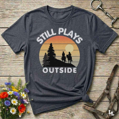 A dark charcoal gray t-shirt with a retro sunset graphic and the text 'STILL PLAYS OUTSIDE' in white. The shirt is styled in a flat-lay on a rustic wooden background with equestrian accessories (bridle) and a small bouquet of wildflowers.