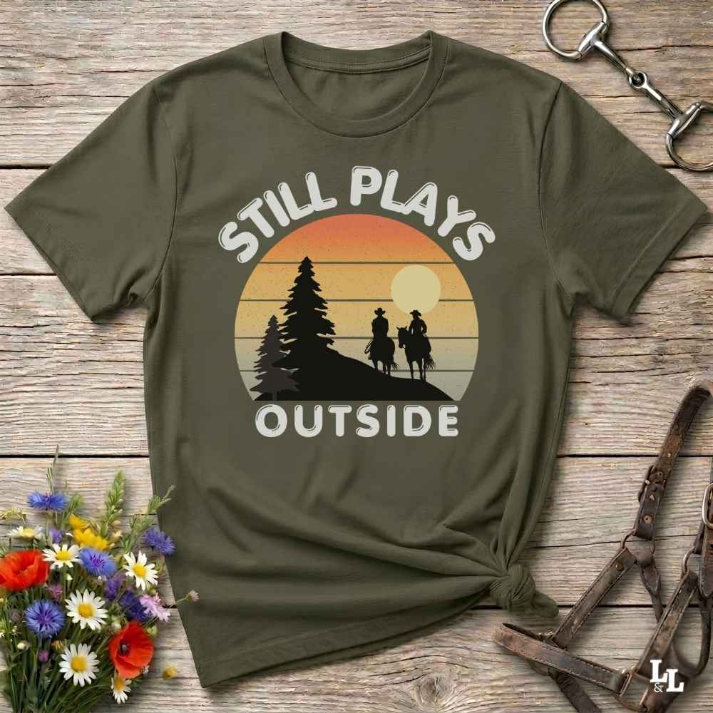 An olive green short-sleeve t-shirt with a retro sunset graphic and the text 'STILL PLAYS OUTSIDE' printed on the front, styled in a flat-lay on a rustic wooden background with flowers and leather accessories.