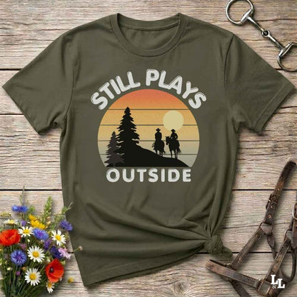 An olive green short-sleeve t-shirt with a retro sunset graphic and the text 'STILL PLAYS OUTSIDE' printed on the front, styled in a flat-lay on a rustic wooden background with flowers and leather accessories.
