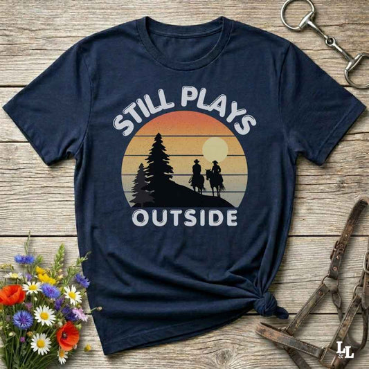 A navy blue graphic t-shirt laid flat on a rustic wooden surface, styled with equestrian accessories and a small bouquet of wildflowers. The t-shirt features a retro sunset graphic with silhouettes of a tree and two riders, with the text 'STILL PLAYS OUTSIDE' in white.