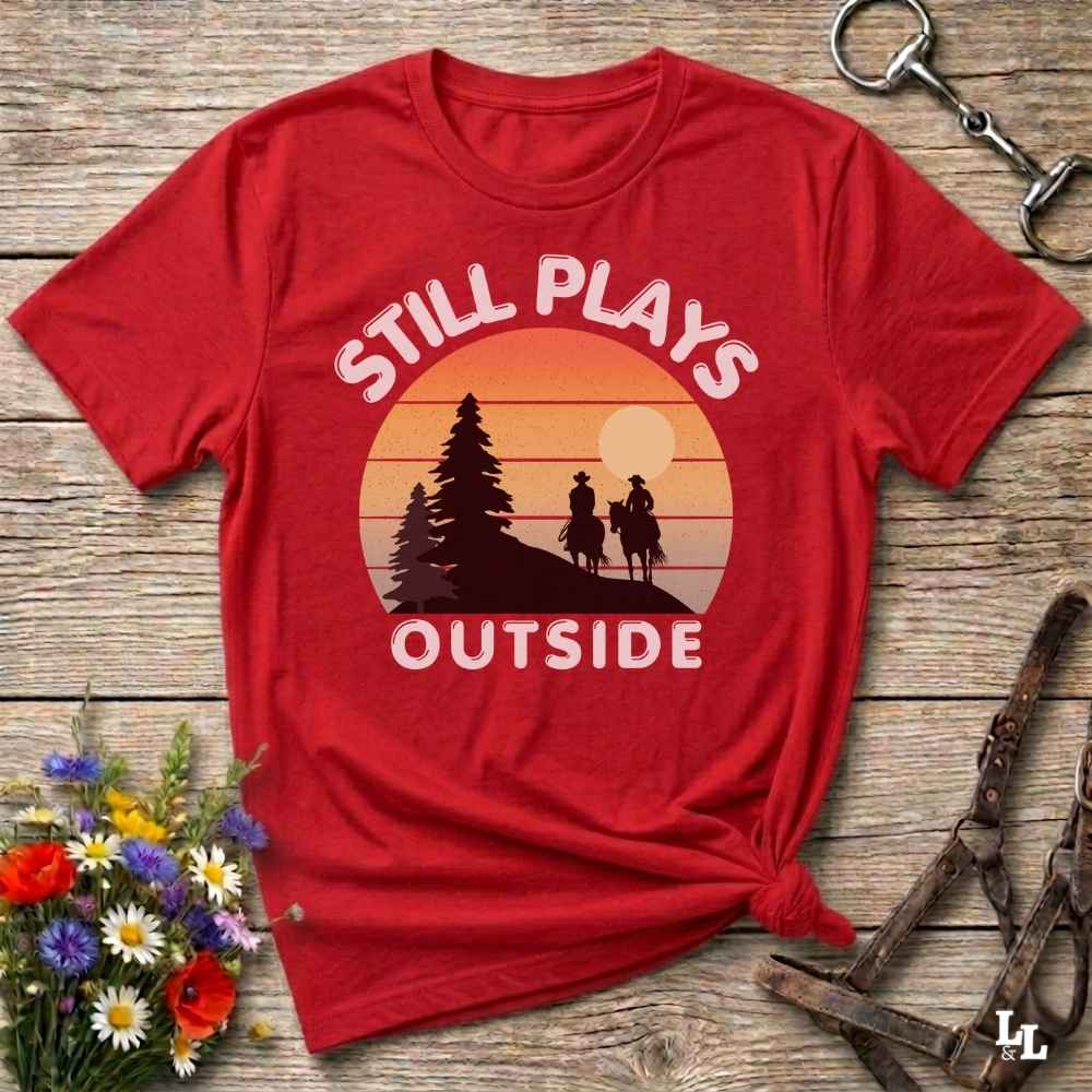 A red short-sleeve graphic T-shirt laid flat on a rustic wooden surface, with a sunset-style print showing two riders on horseback and the words “STILL PLAYS OUTSIDE.” Decorative flowers and horse tack are placed around it as styling props.