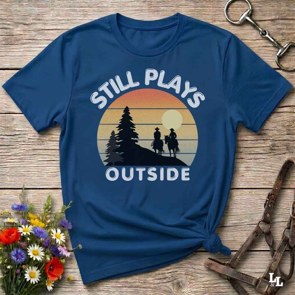 A navy blue graphic t-shirt laid flat on a rustic wooden surface, styled with a small bouquet of wildflowers, a horse bit, and a leather halter. The t-shirt features a retro sunset graphic with silhouettes of a pine tree and two riders on horseback, with the text 'STILL PLAYS OUTSIDE' in white.