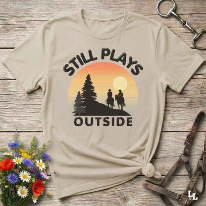 A beige graphic t-shirt with a retro sunset design and the text 'STILL PLAYS OUTSIDE' in black, styled in a flat-lay on a rustic wooden background with wildflowers and leather straps.