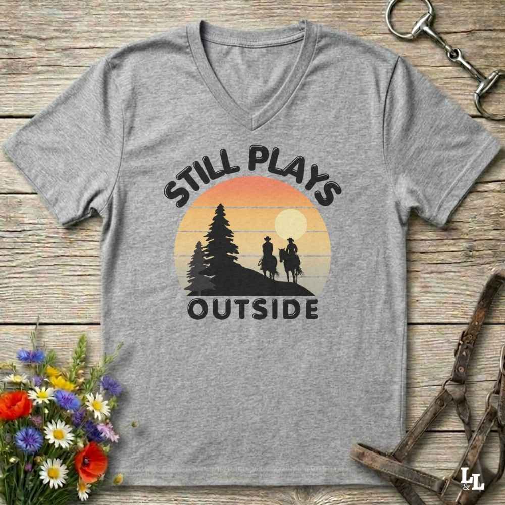 A heather gray V-neck t-shirt with a retro-style sunset graphic and the text 'STILL PLAYS OUTSIDE' printed on the front, displayed flat on a rustic wooden surface with decorative flowers and horse tack props.