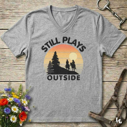 A heather gray V-neck t-shirt with a retro-style sunset graphic and the text 'STILL PLAYS OUTSIDE' printed on the front, displayed flat on a rustic wooden surface with decorative flowers and horse tack props.
