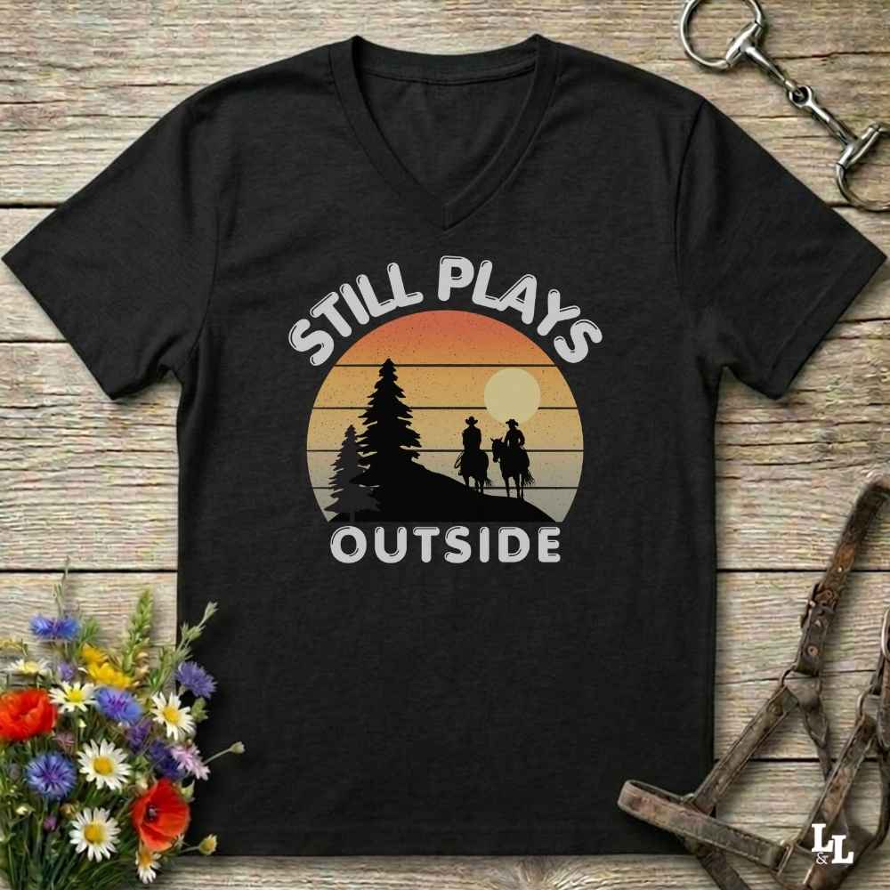 A black V-neck t-shirt with a retro sunset graphic and the text 'STILL PLAYS OUTSIDE' printed on the front, displayed on a rustic wooden background with equestrian accessories and flowers.