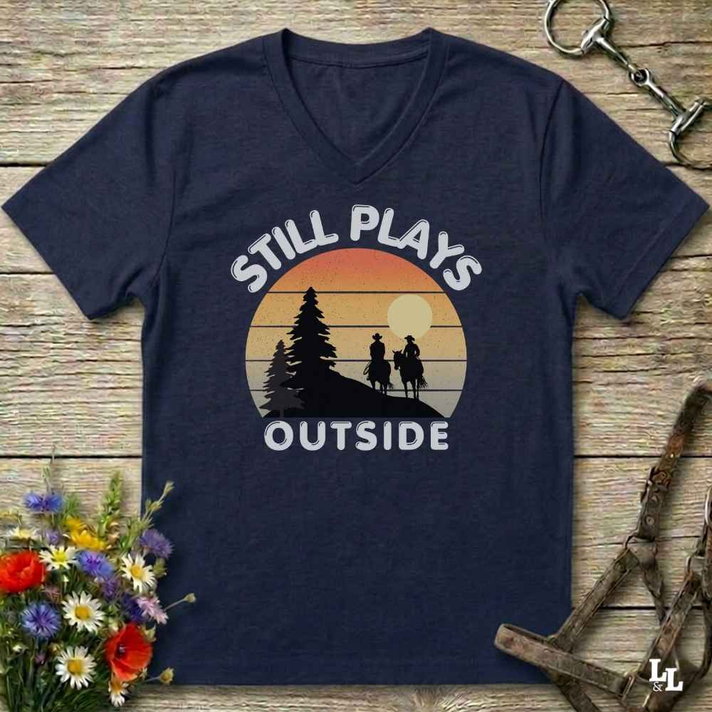 A navy blue V-neck t-shirt laid flat on a rustic wooden surface, featuring a retro sunset graphic with the text 'STILL PLAYS OUTSIDE' and silhouettes of a tree and riders. Decorative flowers and horse tack are placed around the shirt.