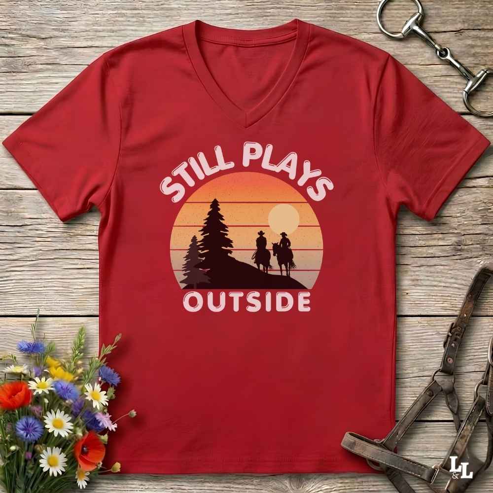 A red V-neck t-shirt laid flat on a rustic wooden surface, featuring a retro sunset graphic with the text 'STILL PLAYS OUTSIDE' and silhouettes of a person riding horses and a pine tree. The shirt is styled with equestrian tack and a small bouquet of wildflowers.