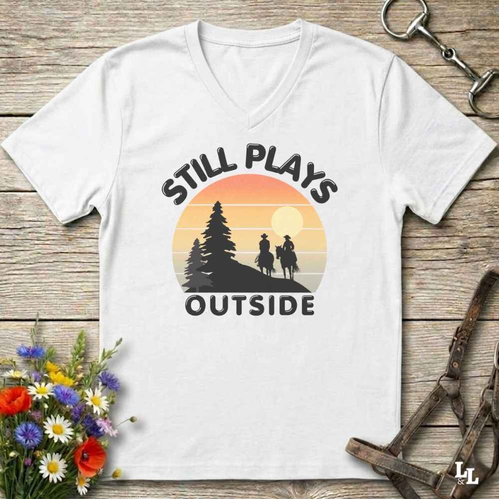 A white V-neck t-shirt with a retro sunset graphic and the text 'STILL PLAYS OUTSIDE' printed on the front, displayed on a rustic wooden background with decorative flowers and tack.