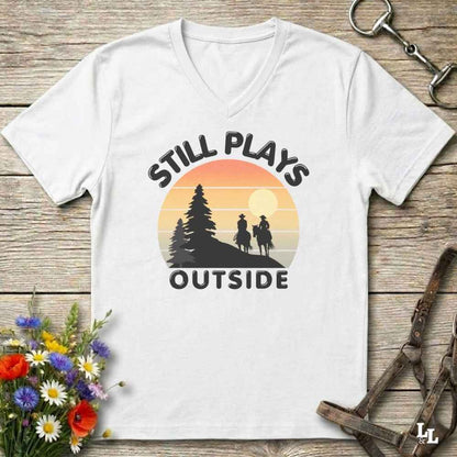 A white V-neck t-shirt with a retro sunset graphic and the text 'STILL PLAYS OUTSIDE' printed on the front, displayed on a rustic wooden background with decorative flowers and tack.