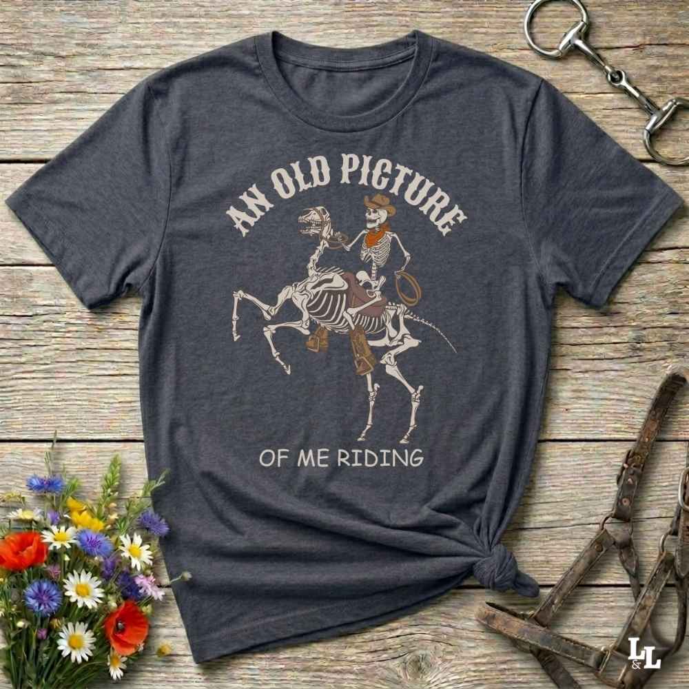 A dark charcoal gray graphic t-shirt laid flat on a rustic wooden surface, styled with a small bouquet of flowers, a horse bit, and a leather halter. The t-shirt features a graphic of a skeleton cowboy riding a skeleton horse, with the text 'AN OLD PICTURE OF ME RIDING'.