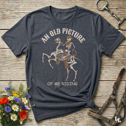 A dark charcoal gray graphic t-shirt laid flat on a rustic wooden surface, styled with a small bouquet of flowers, a horse bit, and a leather halter. The t-shirt features a graphic of a skeleton cowboy riding a skeleton horse, with the text 'AN OLD PICTURE OF ME RIDING'.
