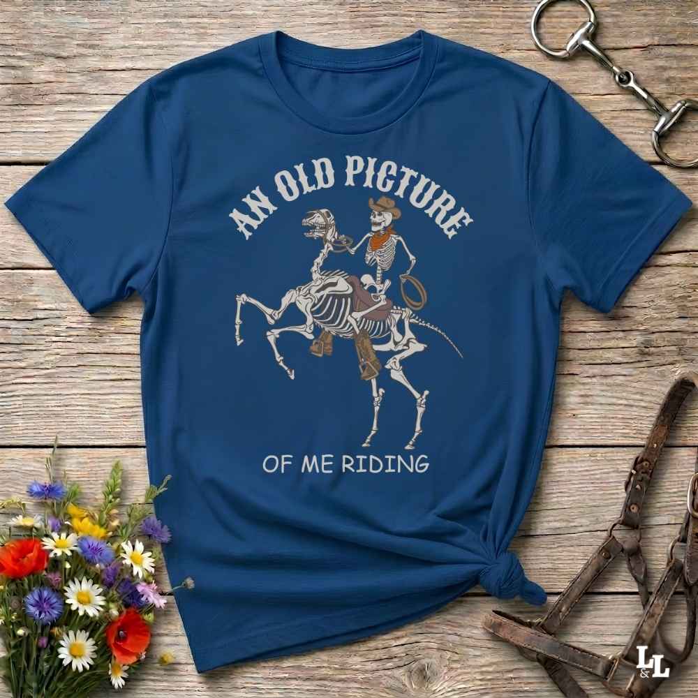 A navy blue graphic t-shirt laid flat on a rustic wooden surface, styled with a small bouquet of flowers, a horse bit, and leather reins. The shirt features a graphic of a skeleton rider on a horse skeleton with the text 'AN OLD PICTURE OF ME RIDING'.
