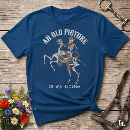 A navy blue graphic t-shirt laid flat on a rustic wooden surface, styled with a small bouquet of flowers, a horse bit, and leather reins. The shirt features a graphic of a skeleton rider on a horse skeleton with the text 'AN OLD PICTURE OF ME RIDING'.