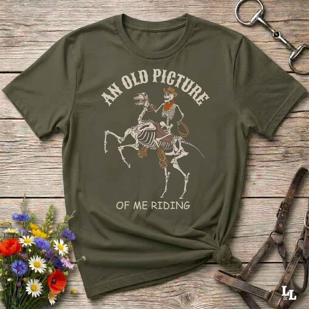 An olive green graphic t-shirt laid flat on a rustic wooden surface, styled with a horse bit, leather reins, and a small bouquet of colorful wildflowers. The t-shirt features a graphic of a skeleton cowboy riding a skeleton horse, with the text 'AN OLD PICTURE OF ME RIDING'.