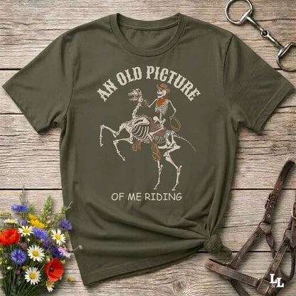 An olive green graphic t-shirt laid flat on a rustic wooden surface, styled with a horse bit, leather reins, and a small bouquet of colorful wildflowers. The t-shirt features a graphic of a skeleton cowboy riding a skeleton horse, with the text 'AN OLD PICTURE OF ME RIDING'.
