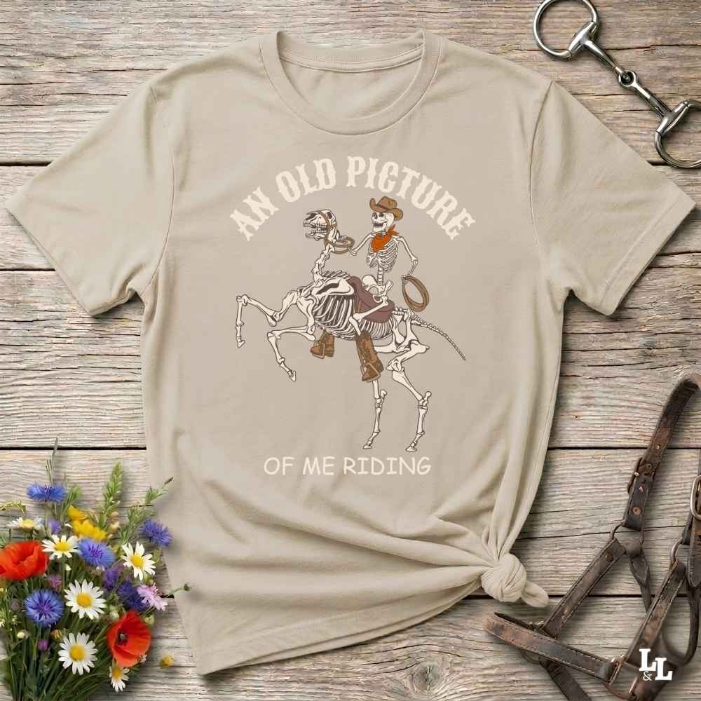 A beige graphic t-shirt laid flat on a rustic wooden surface, styled with flowers, a horse bit, and a leather bridle. The shirt features a western skeleton cowboy riding a skeleton horse with the text 'AN OLD PICTURE OF ME RIDING'.