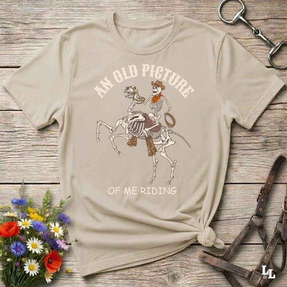 A beige graphic t-shirt laid flat on a rustic wooden surface, styled with flowers, a horse bit, and a leather bridle. The shirt features a western skeleton cowboy riding a skeleton horse with the text 'AN OLD PICTURE OF ME RIDING'.