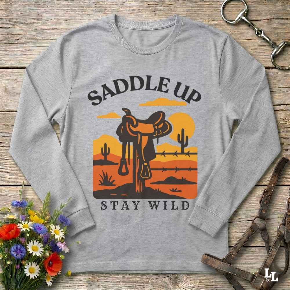A light heather gray long-sleeve t-shirt with a western-themed graphic print reading 'SADDLE UP' and 'STAY WILD'. The graphic features a brown leather saddle in front of a desert sunset with cacti.