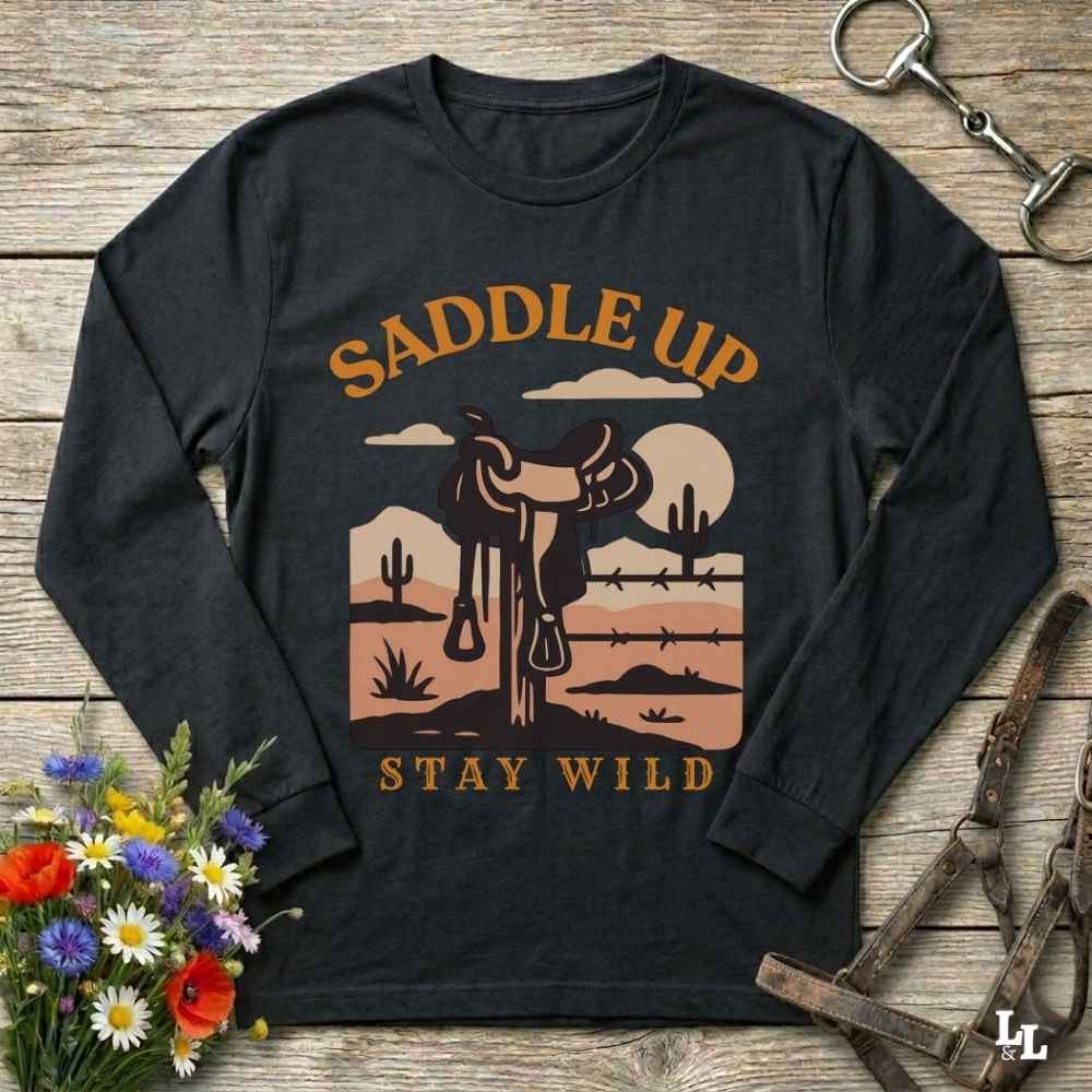 A black long-sleeve t-shirt with a western-themed graphic print reading 'SADDLE UP' and 'STAY WILD'. The graphic features a cowboy silhouette, a saddle, cacti, and a sunset landscape in warm brown, orange, and beige tones.