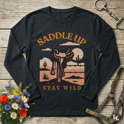 A black long-sleeve t-shirt with a western-themed graphic print reading 'SADDLE UP' and 'STAY WILD'. The graphic features a cowboy silhouette, a saddle, cacti, and a sunset landscape in warm brown, orange, and beige tones.
