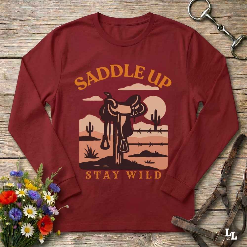 A long-sleeve crewneck t-shirt in maroon red, laid flat on a rustic wooden background. The shirt features a western-themed graphic with a saddle, desert landscape, and the text 'SADDLE UP' and 'STAY WILD' in orange and beige.
