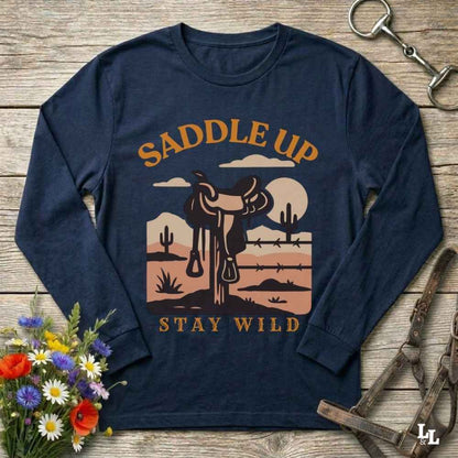 A navy blue long-sleeve t-shirt with a western-themed graphic print featuring a saddle, desert landscape, and the text 'SADDLE UP' and 'STAY WILD'. The shirt is displayed flat on a rustic wooden background with equestrian accessories and flowers.