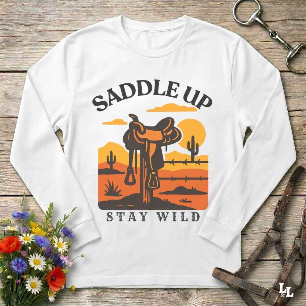 A white long-sleeve crewneck t-shirt with a western-themed graphic print. The graphic features a brown saddle in a desert landscape with cacti, a yellow sun, and the text 'SADDLE UP' above and 'STAY WILD' below.