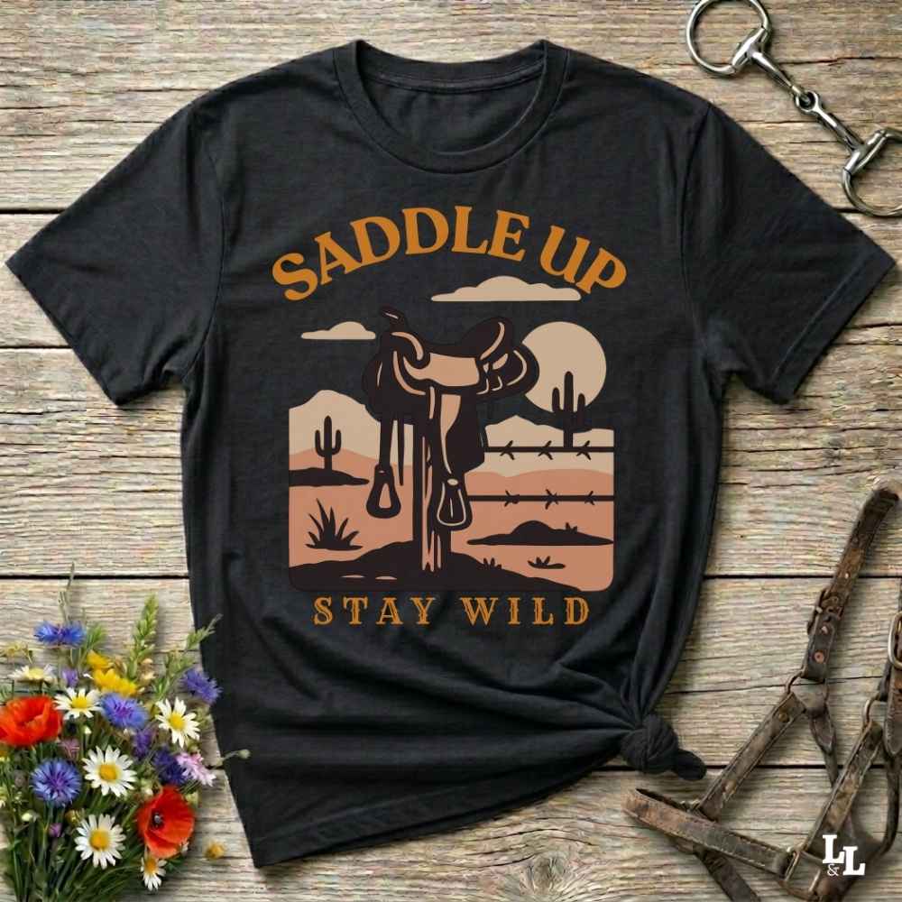 A black graphic t-shirt laid flat on a rustic wooden surface, styled with a halter, flowers, and a horse bit. The shirt features a western desert landscape graphic with a steer, cacti, and the text 'SADDLE UP' and 'STAY WILD' in tan and orange.