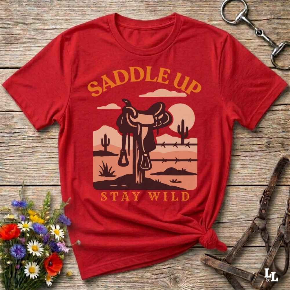 A red short-sleeve graphic T-shirt laid flat on a rustic wood surface, with a western desert scene printed in tan and brown. The shirt reads “SADDLE UP” at the top and “STAY WILD” at the bottom.