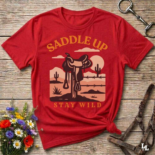 A red short-sleeve graphic T-shirt laid flat on a rustic wood surface, with a western desert scene printed in tan and brown. The shirt reads “SADDLE UP” at the top and “STAY WILD” at the bottom.