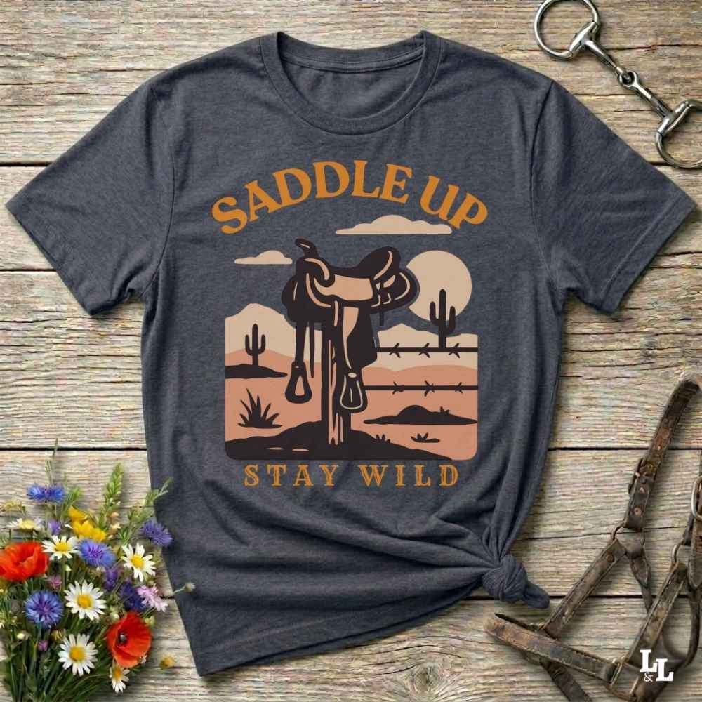 A dark heather gray graphic t-shirt laid flat on a rustic wooden background, styled with a western saddle, horse bits, and a small bouquet of wildflowers. The shirt features a graphic of a western saddle in a desert landscape with the text 'SADDLE UP' and 'STAY WILD'.