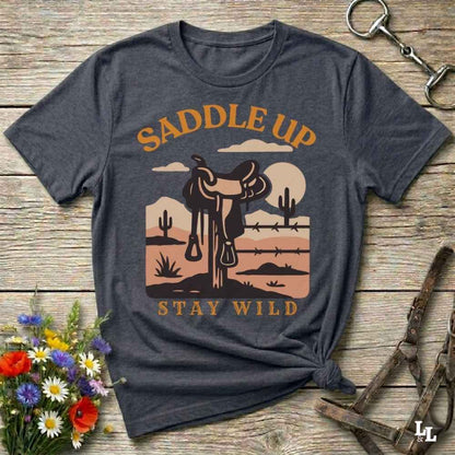 A dark heather gray graphic t-shirt laid flat on a rustic wooden background, styled with a western saddle, horse bits, and a small bouquet of wildflowers. The shirt features a graphic of a western saddle in a desert landscape with the text 'SADDLE UP' and 'STAY WILD'.