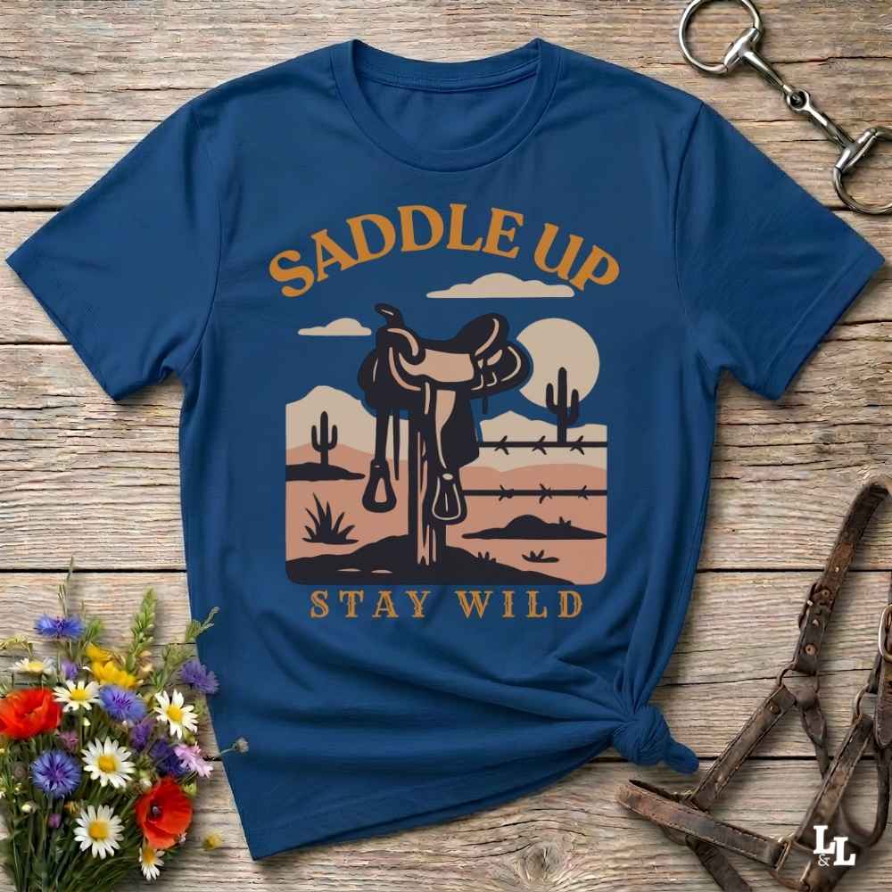 A navy blue graphic t-shirt laid flat on a rustic wooden surface, styled with equestrian and floral props. The shirt features a retro desert landscape graphic with a saddle, cactus, and barbed wire, framed by the words 'SADDLE UP' and 'STAY WILD' in tan lettering.