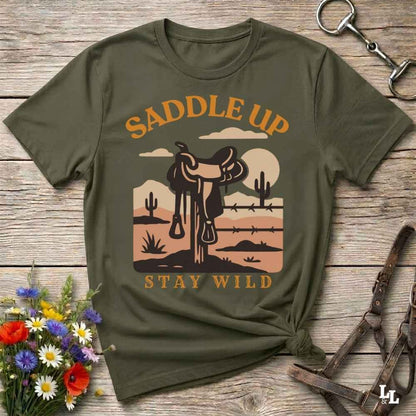 An olive green graphic t-shirt laid flat on a rustic wooden surface, styled with a western saddle design and the words 'SADDLE UP' and 'STAY WILD'.