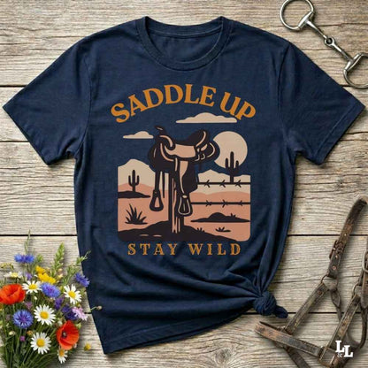 A navy blue graphic t-shirt with a western theme, laid flat on a rustic wooden background with decorative horse tack and flowers. The shirt features a graphic of a saddle on a desert landscape with the text 'SADDLE UP' and 'STAY WILD'.