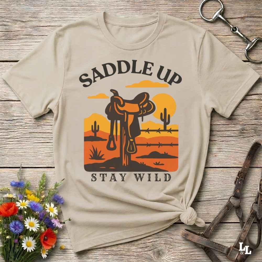 A beige graphic t-shirt laid flat on a rustic wooden surface, styled with equestrian accessories and a small bouquet of flowers. The shirt features a western desert landscape graphic with a saddle, cacti, and the text 'SADDLE UP' and 'STAY WILD'.