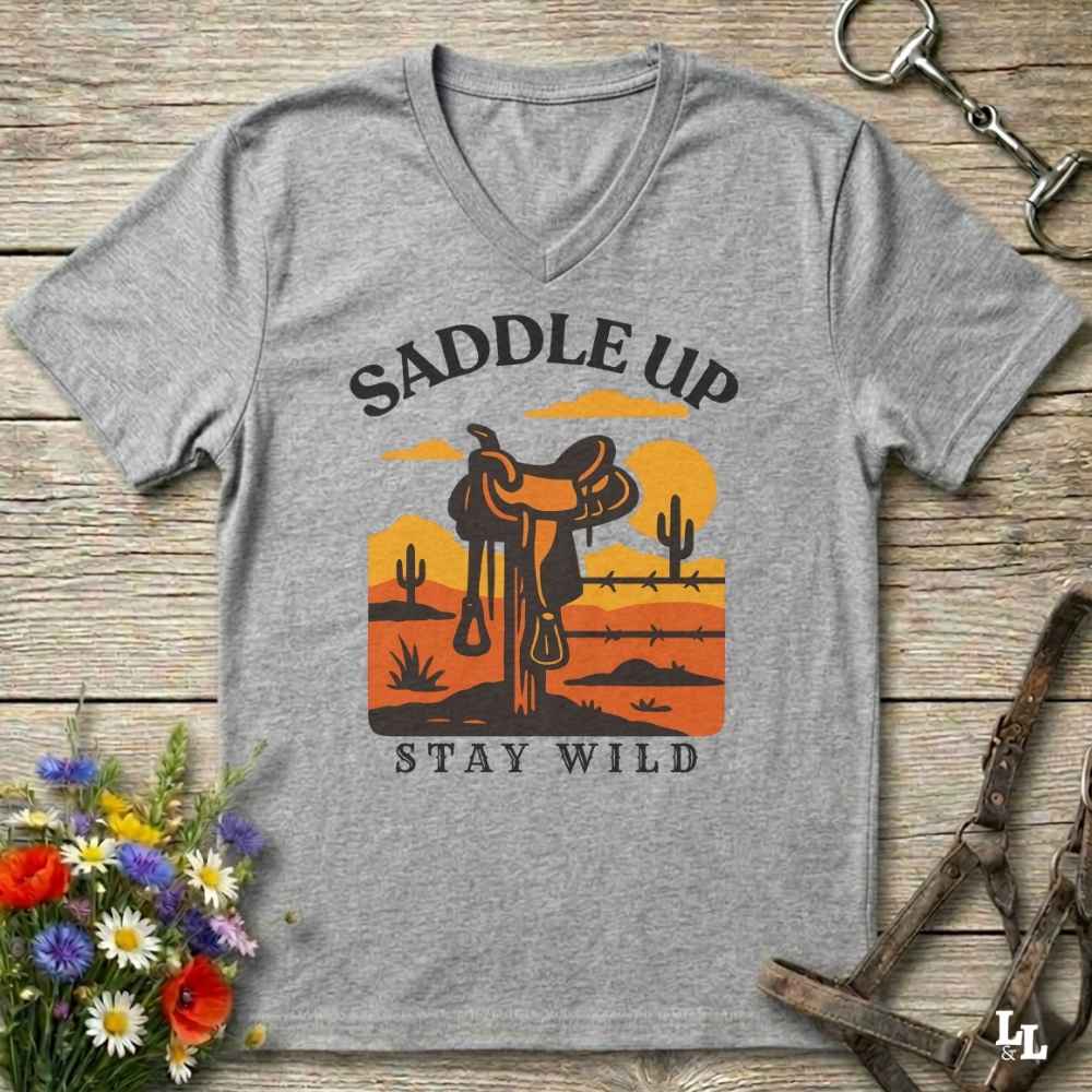 A heather gray V-neck t-shirt with a western-themed graphic print reading 'SADDLE UP' and 'STAY WILD'. The graphic features a cowboy saddle in front of a desert sunset with cacti.