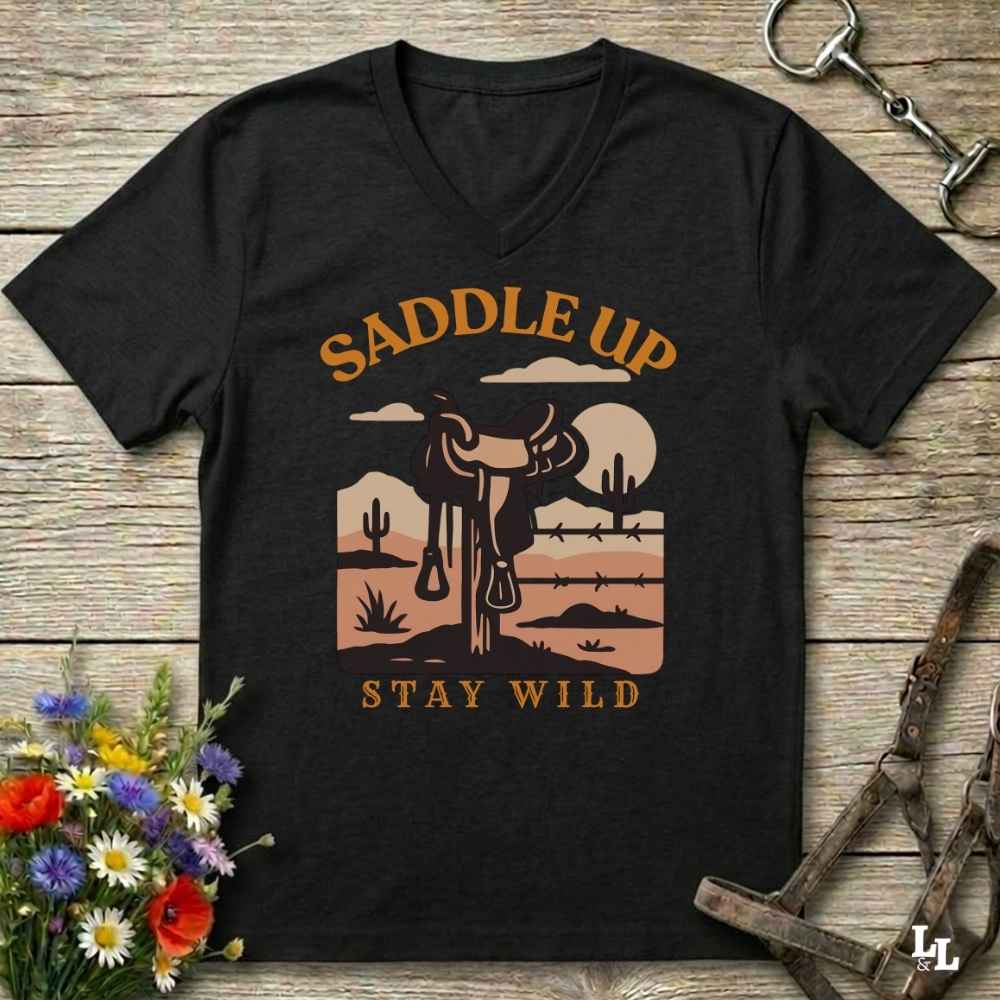 A black V-neck t-shirt with a retro western desert graphic featuring a saddle, cacti, and the text 'SADDLE UP STAY WILD' in tan and orange, laid flat on a rustic wooden background with horse tack and flowers as props.