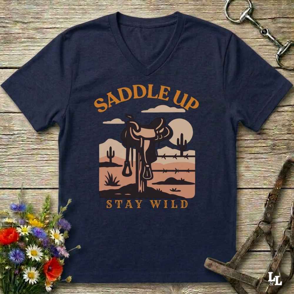A navy blue V-neck t-shirt with a retro western graphic that reads 'SADDLE UP' and 'STAY WILD' around an illustration of a saddle, desert landscape, and sun. The shirt is displayed flat on a rustic wooden background with decorative horse tack and flowers.