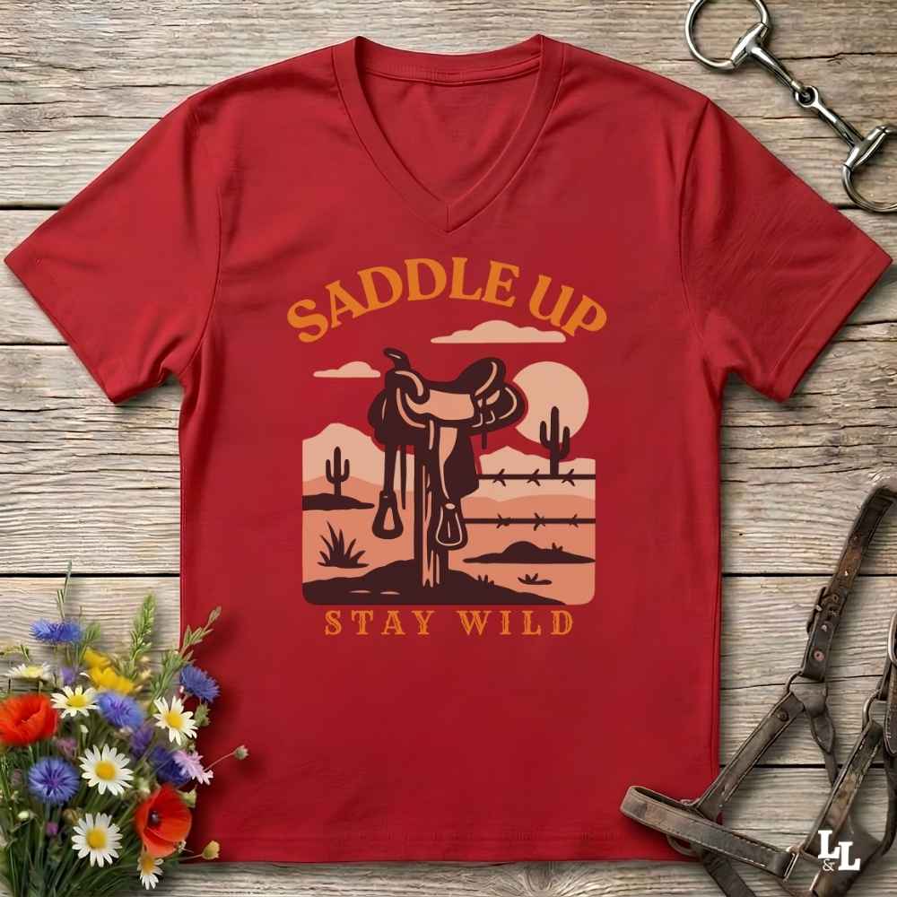 A red V-neck t-shirt with a western-themed desert landscape graphic and the text 'SADDLE UP STAY WILD' in orange and beige, laid flat on a rustic wooden background with horse tack and flowers as props.