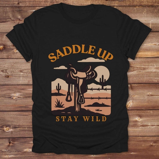 Saddle Up Stay Wild western t-shirt features a bold desert sunset graphic with a classic saddle and western landscape. Perfect for cowgirls, cowboys, and horse lovers, this rustic tee blends adventure with comfort and country charm. Whether you're heading to a rodeo, trail ride, or simply embracing your wild side, this shirt offers a soft fit and bold design. Designed for western wear fans, equestrians, and country souls, it's a must have for anyone who lives the saddle life. 