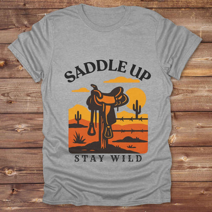 Saddle Up Stay Wild western t-shirt features a bold desert sunset graphic with a classic saddle and western landscape. Perfect for cowgirls, cowboys, and horse lovers, this rustic tee blends adventure with comfort and country charm. Whether you're heading to a rodeo, trail ride, or simply embracing your wild side, this shirt offers a soft fit and bold design. Designed for western wear fans, equestrians, and country souls, it's a must have for anyone who lives the saddle life. 