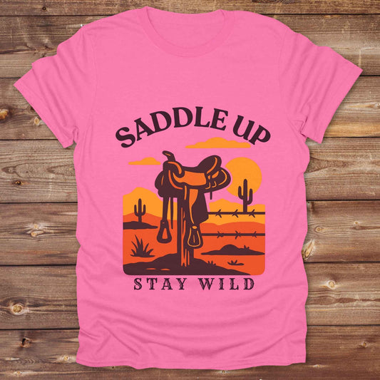 Saddle Up Stay Wild western t-shirt features a bold desert sunset graphic with a classic saddle and western landscape. Perfect for cowgirls, cowboys, and horse lovers, this rustic tee blends adventure with comfort and country charm. Whether you're heading to a rodeo, trail ride, or simply embracing your wild side, this shirt offers a soft fit and bold design. Designed for western wear fans, equestrians, and country souls, it's a must have for anyone who lives the saddle life. 