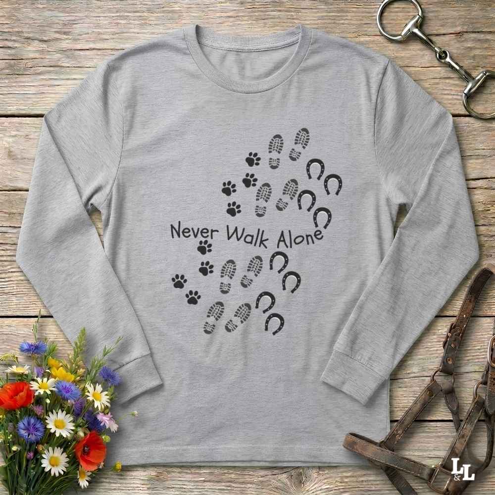 A light gray long-sleeve crewneck shirt laid flat on a rustic wooden surface, featuring a black graphic of paw prints, hoof prints, and the text 'Never Walk Alone.' Decorative flowers and equestrian tack are placed around the shirt.