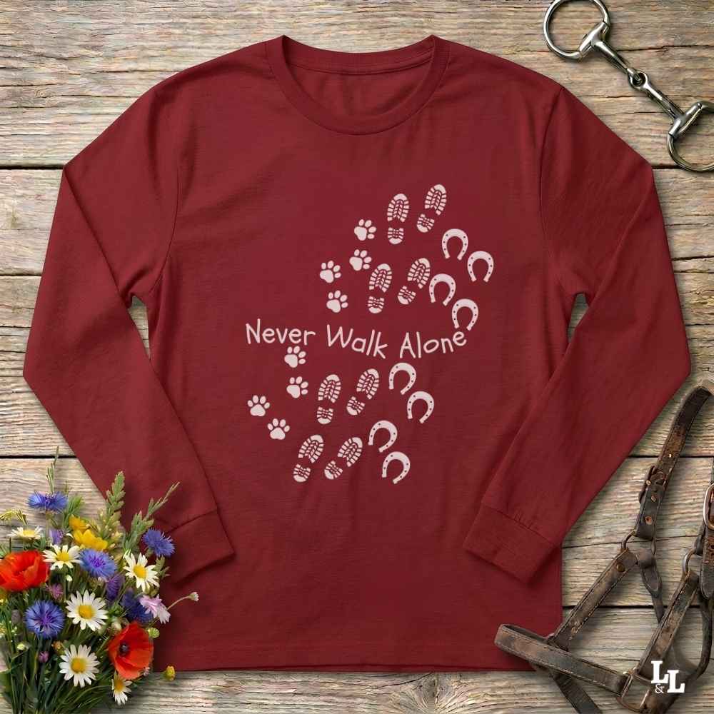 A long-sleeve crewneck t-shirt in maroon red, laid flat on a rustic wooden surface. The shirt features a white graphic print of horse hooves and paw prints arranged in a heart shape around the text 'Never Walk Alone'.